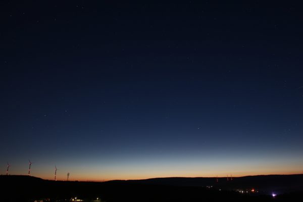 Foto eines Sternenhimmels bei Nacht. Im Hintergrund sieht man die letzten Strahlen der Sonne am Horizont verschwinden. Das dunkle blau des Nachthimmels übernimmt und die letzten Reste der orangenen Färbung verschwindet im Hintergrund.
